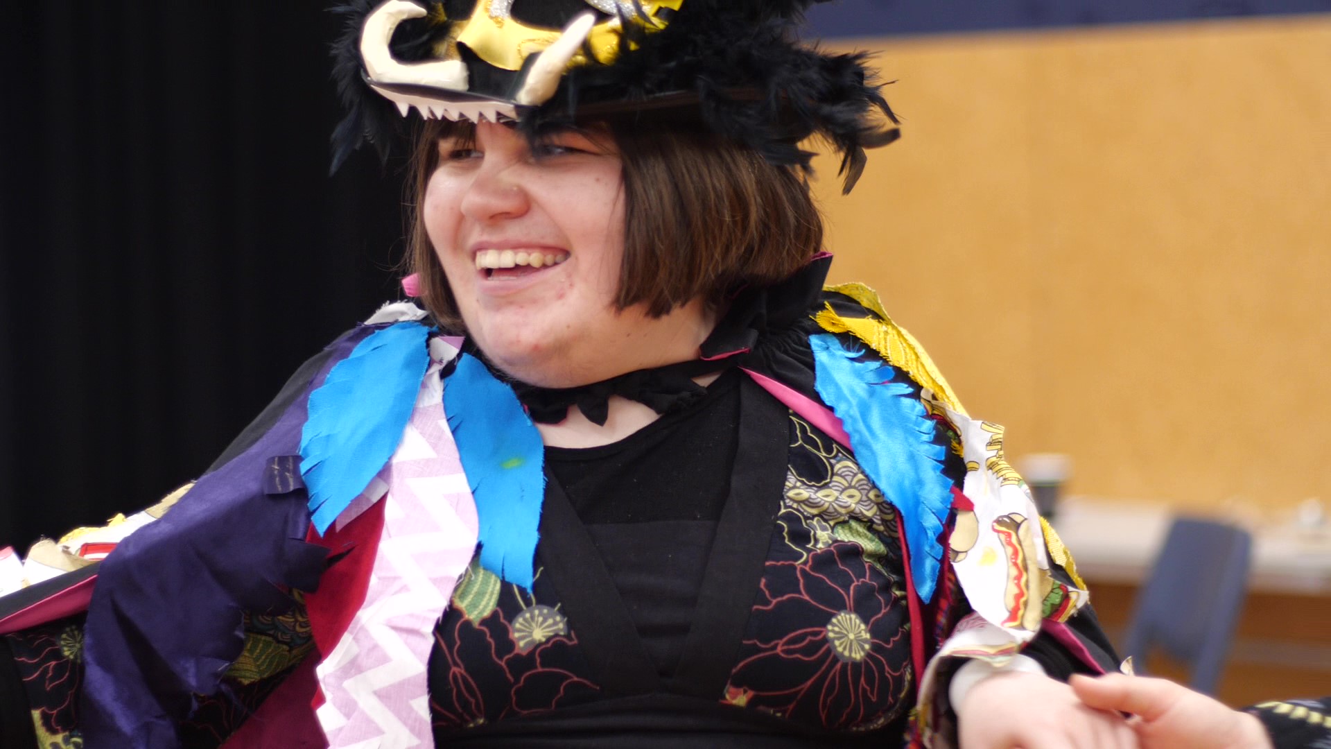 A close up of Maggie Rose Paine showing her Mag Bird Wearable Arts piece at the Disability Pride Week celebration at Te Whaea – National Dance and Drama Centre, Wellington, December 2018. A Mag bird is a cross between a Kunikuni pig and a staunch eagle. Maggie Rose is wearing a black top hat. The hat is covered in fluffy black feathers and has two cream coloured clay tusks on the front brim. She has a big smile on her face and looks like she is in her element! Her Mag Bird cloak is draped over her shoulders. It is covered in feathers made of all different sorts of patterned and colourful cloth.