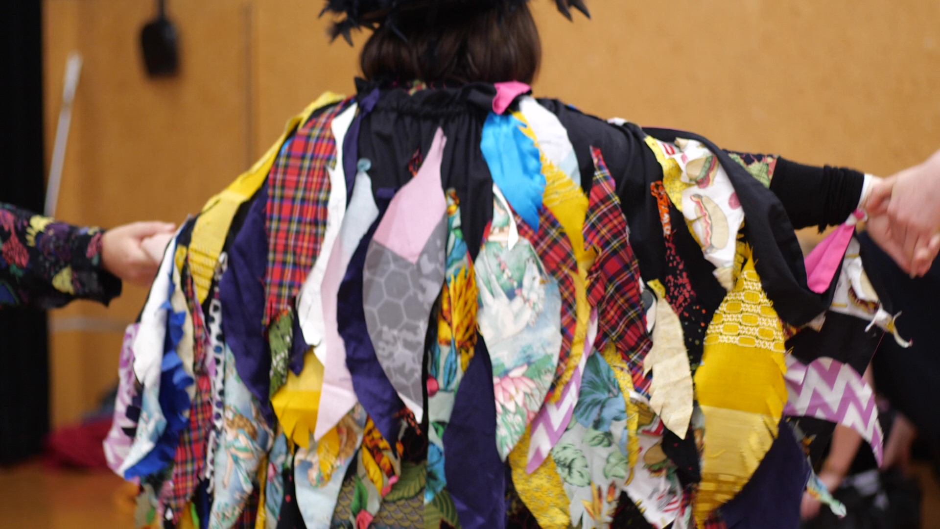 In this image Mag Bird has her arms extended to show the back of her rainbow feather cloak and wings. She is holding hands with her team, one on each side, as she extends her wings. Her cloak is covered in dozens of feathers that were cut from cloth and sewn onto a cape. The feathers are layered and hanging down. There are lots of different patterns and bright colours. The tartan feathers are Maggie’s favourite. Maggie describes the Mag Bird as having “rainbow feathers”.