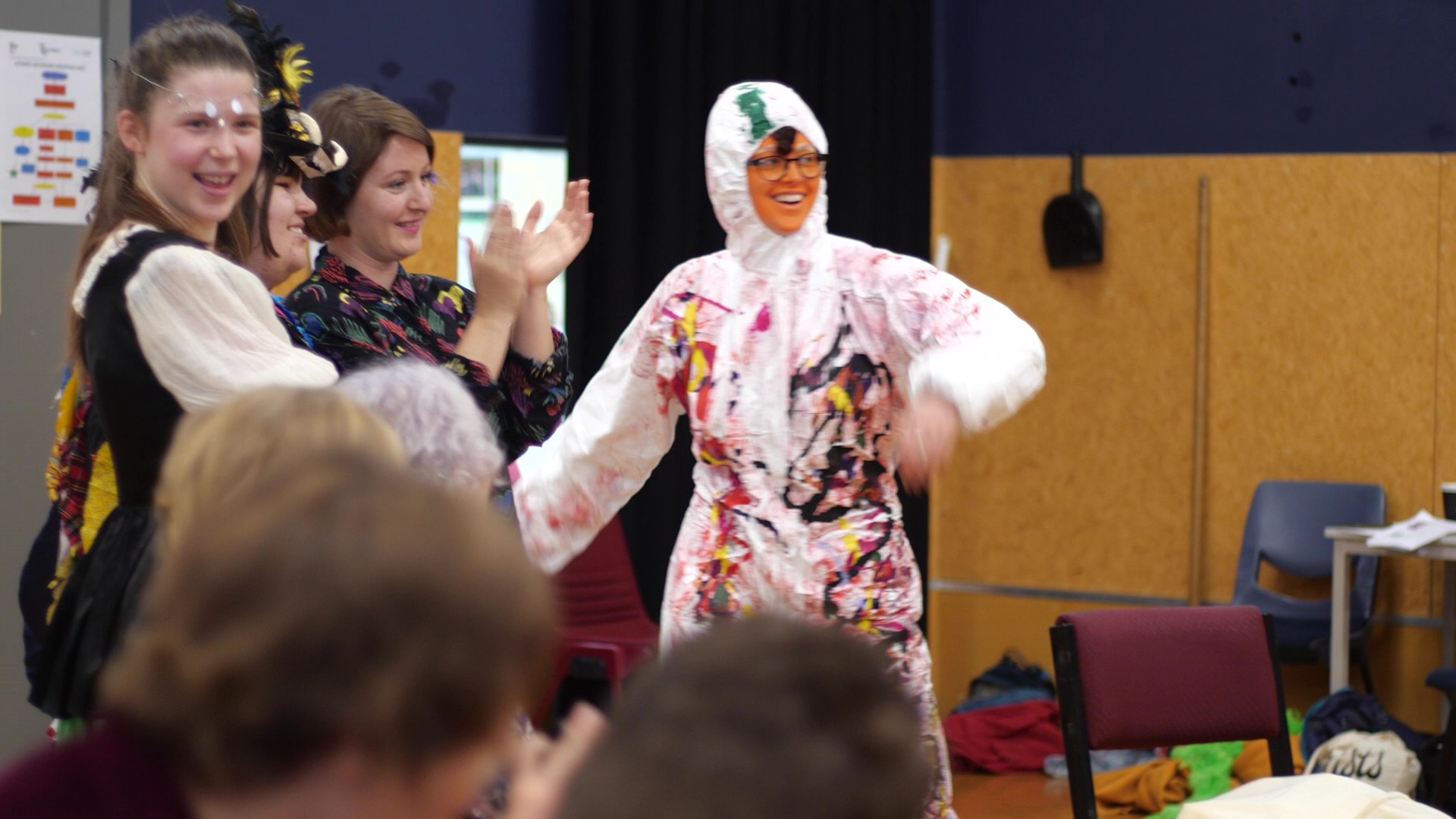 This image is of Paul Holme’s Disabled Oompah Loompah and Maggie Paine’s Mag Bird standing with the Mag Bird’s team Bethany and Sabrina. Sabrina is smiling and clapping. They are looking at something happening out of view of camera view. The Oompah Loompah is laughing, holding one hand on her hip and flapping her other arm.