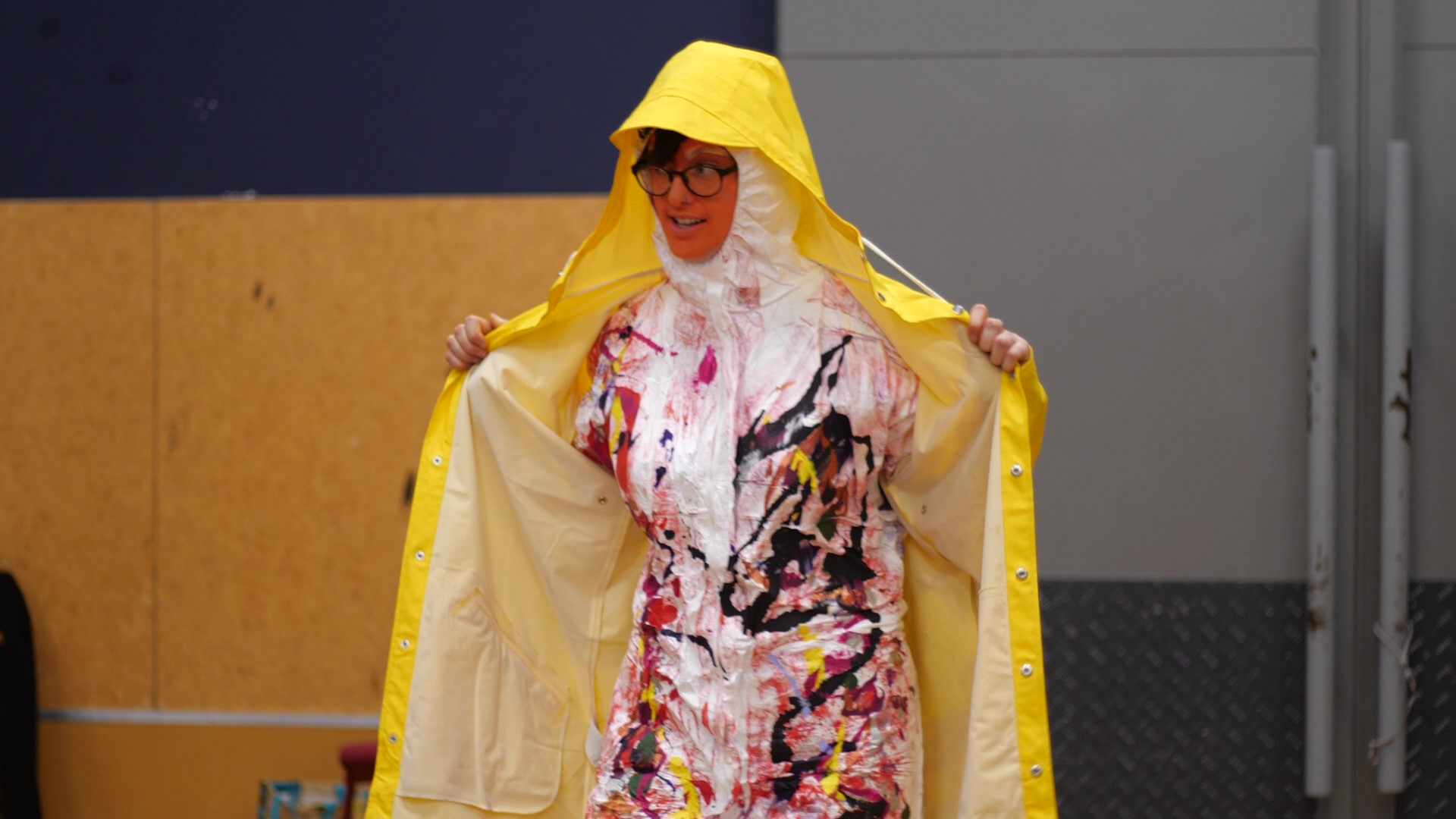 This image is of Sylvie McCreanor modelling Paul Holme’s wearable arts piece, a Disabled Oompah Loompah. Her face is painted bright orange, she is smiling and looking to the audience at her side. She is wearing black rimmed eye glasses. Sylvie is wearing a bright yellow raincoat over brightly spray painted white paper overalls. The hoods for the overalls and coat are up. She is holding the raincoat wide open to expose the overalls underneath. Paul has drizzled and splattered black, yellow, orange, red, purple and pink paint down the front of the overalls. The pattern looks like a multi-coloured, braided swirling river and continues from shoulders to feet. The back of the overalls are spray painted purple and the hood has a rough stripe of green. The Ooompah Loompah music from Charlie and the Chocolate Factory is playing.